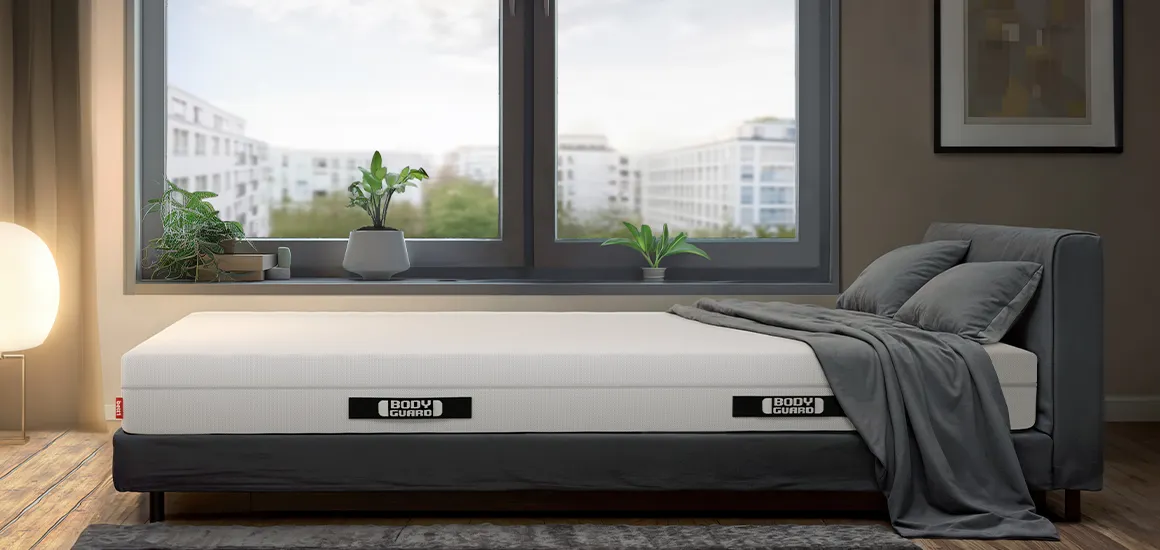 Colchón BODYGUARD® sobre una cama gris en un dormitorio moderno con gran ventana y vistas a la ciudad.