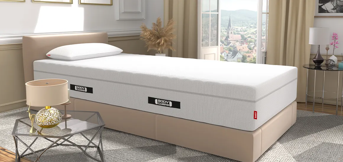 Die BODYGUARD Matratze auf einem Bett in einem hellen Schlafzimmer. Auf der Matratze liegt das BODYGUARD Visco-Kissen. Im Hintergrund offenstehende, bodentiefe Fenster mit Blick auf eine Stadt, vorne ein Beistelltisch mit einer Lampe.