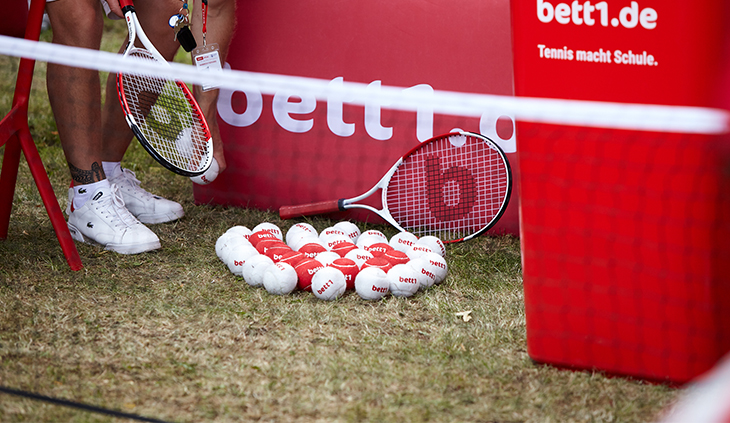 Foto: Hinter einem Tennisnetz liegen rote und weiße Tennisbälle, ein Tennisschläger mit einem b und eine rote Tonne mit der Aufschrift "bett1.de Tennis macht Schule".
