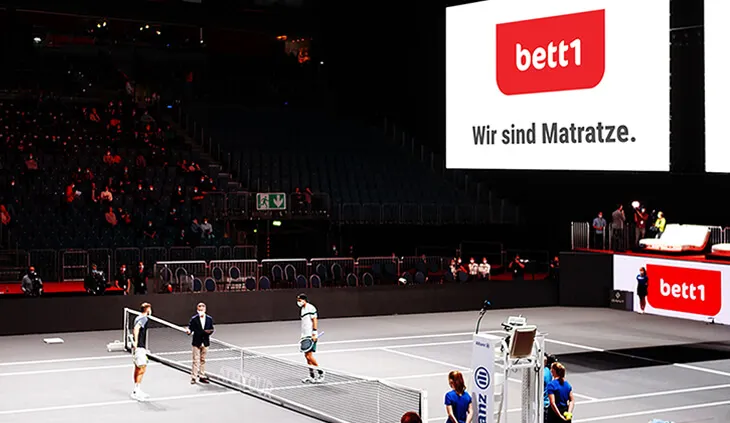 Imagen de una cancha de tenis durante un torneo nocturno patrocinado por bett1.es, mostrando el logotipo en pantallas publicitarias alrededor de la cancha.