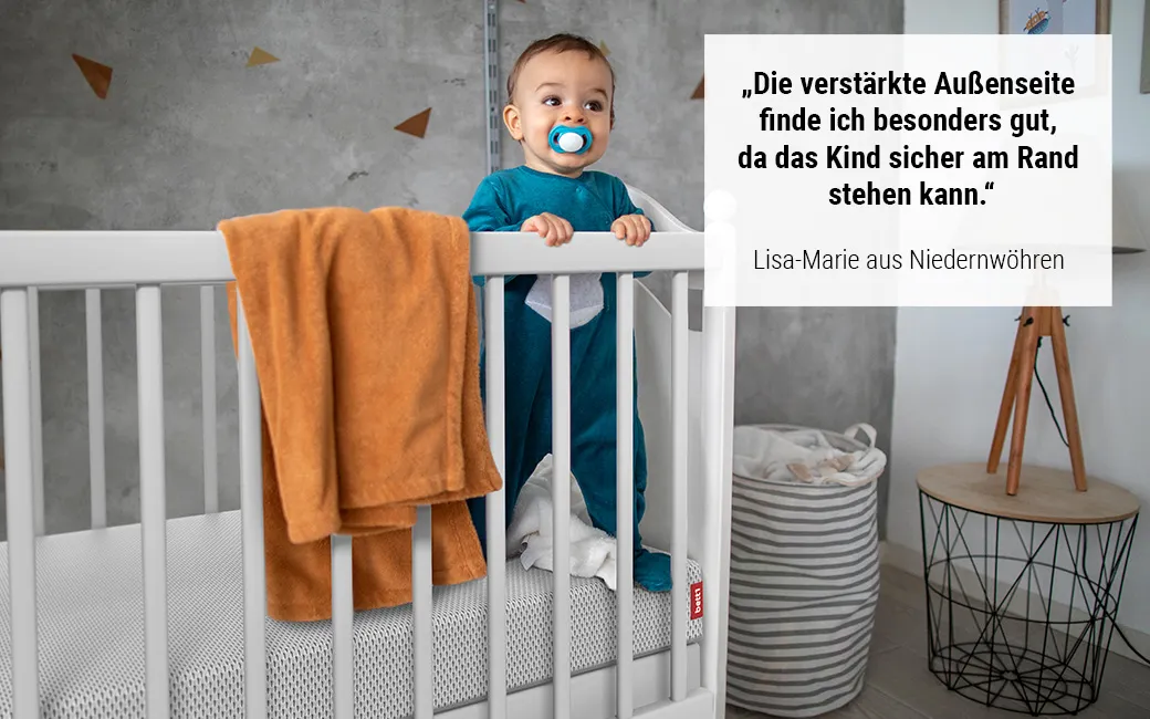 Baby steht in Gitterbett auf der BODYGUARD® Kindermatratze. Text: „Die verstärkte Außenseite finde ich besonders gut, da das Kind sicher am Rand stehen kann.“ Lisa-Marie aus Niedernwöhren	