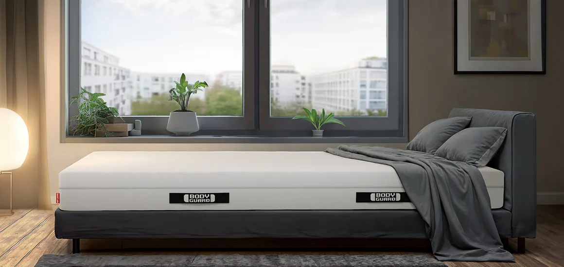 Colchón BODYGUARD® sobre una cama gris en un dormitorio moderno con gran ventana y vistas a la ciudad.