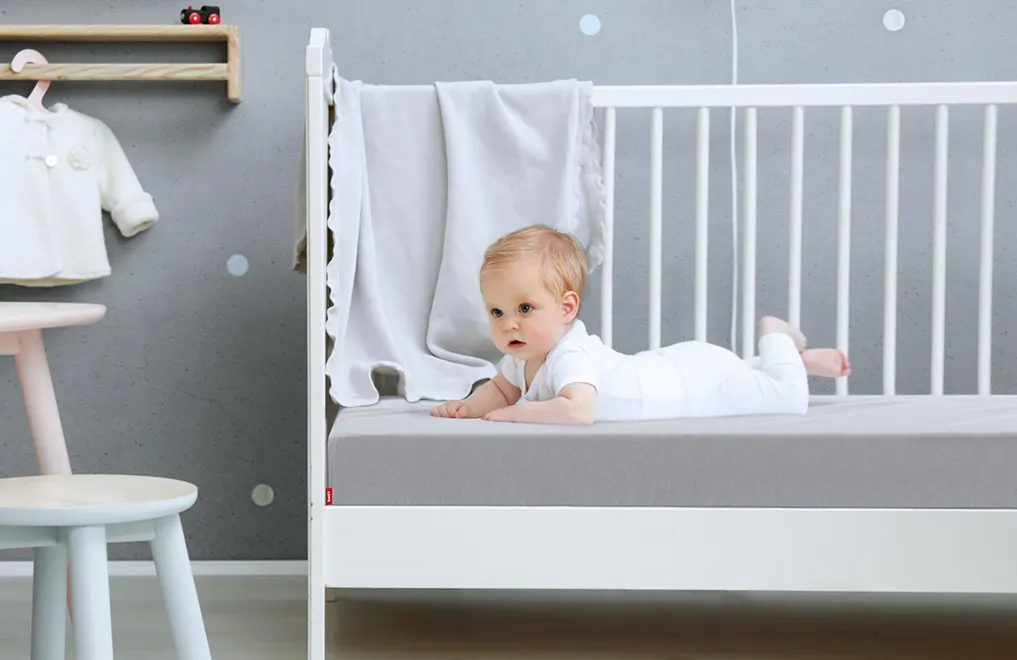 Un bebé acostado en una cuna sobre un colchón de bebé cubierto con una sábana ajustable para niños de color plateado.