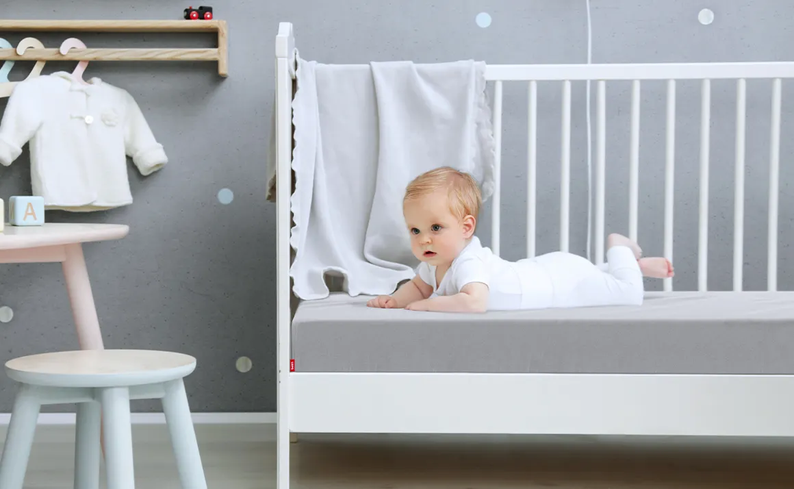 Un bebé acostado en una cuna sobre un colchón de bebé cubierto con una sábana ajustable para niños de color plateado.