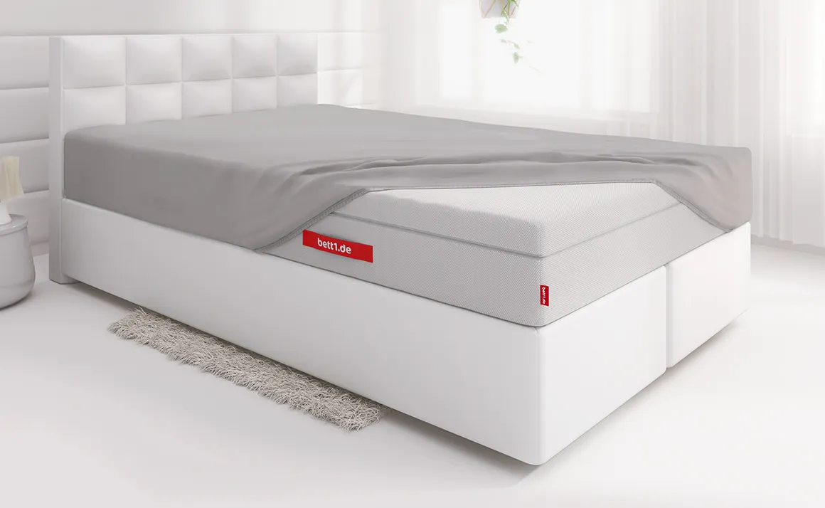Die FLIP® Matratze ist bis auf eine Ecke mit dem Spannbettlaken extra hoch bezogen und liegt auf einem Boxspringbett in einem hellen Schlafzimmer	