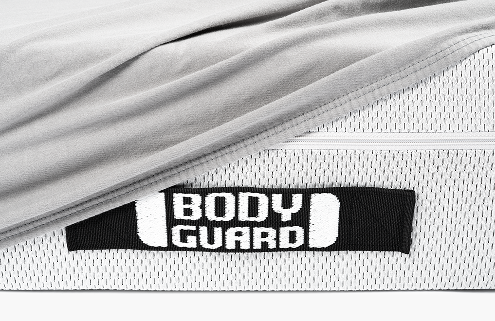 Detalle del colchón Bodyguard con etiqueta de tela, parcialmente cubierto por sábana gris.