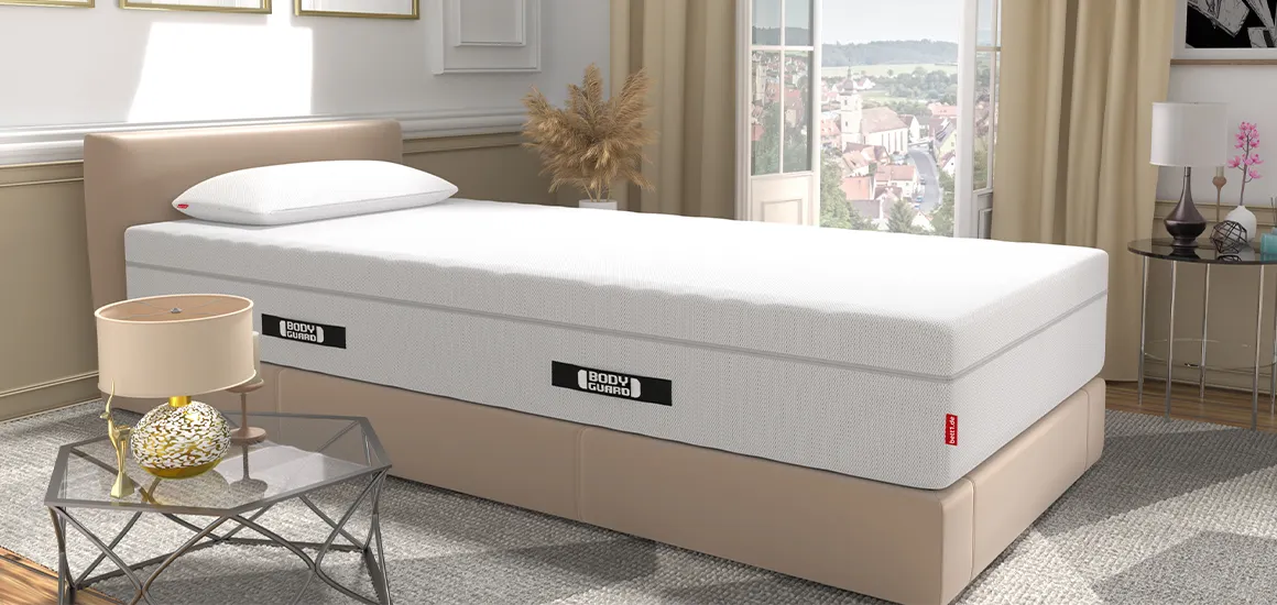 Die BODYGUARD Matratze auf einem Bett in einem hellen Schlafzimmer. Auf der Matratze liegt das BODYGUARD Visco-Kissen. Im Hintergrund offenstehende, bodentiefe Fenster mit Blick auf eine Stadt, vorne ein Beistelltisch mit einer Lampe.